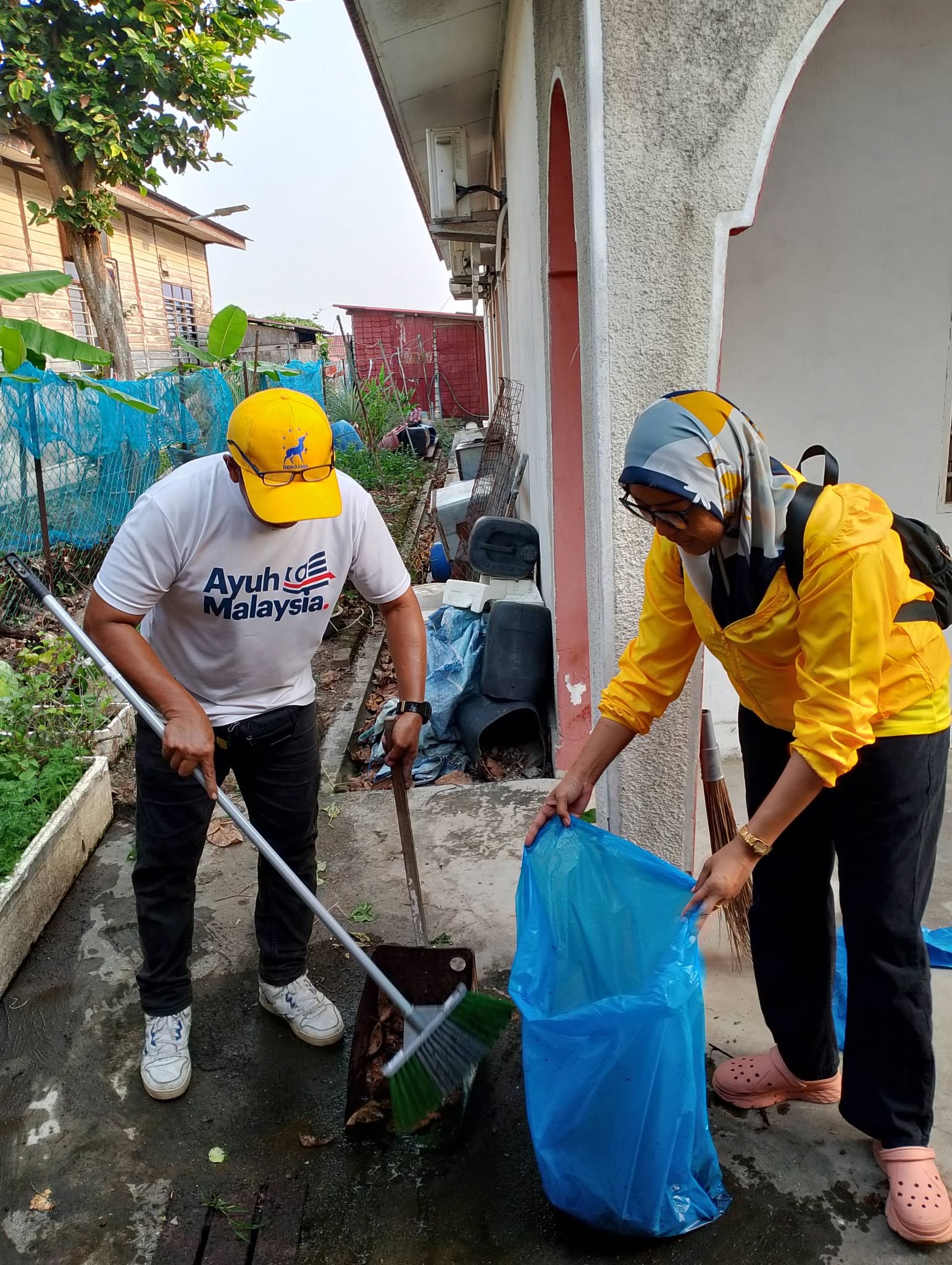 Dua ahli pasukan bekerjasama menyapu dan mengumpulkan sampah ke dalam beg biru.